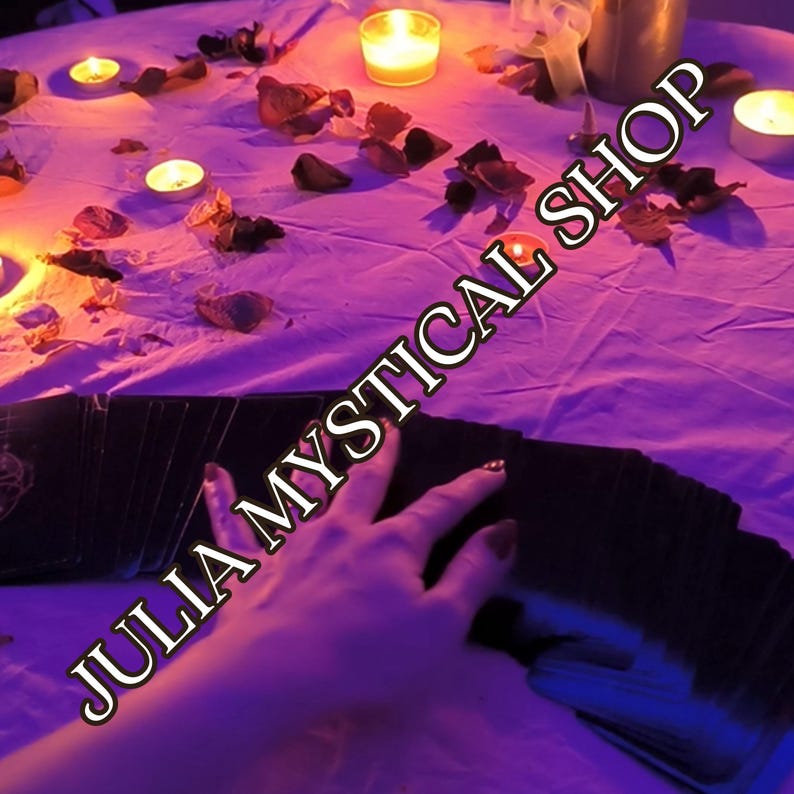 May include: A deck of black tarot cards spread out on a white cloth surface illuminated by purple lighting and several small lit candles. Scattered dried flower petals surround the cards, and the text JULIA MYSTICAL SHOP is overlaid in white.
