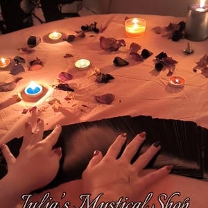 May include: A deck of black tarot cards spread out on a peach-colored surface, surrounded by scattered dried flower petals and several lit tea light candles. The text Julia's Mystical Shop is printed in a white script font at the bottom.