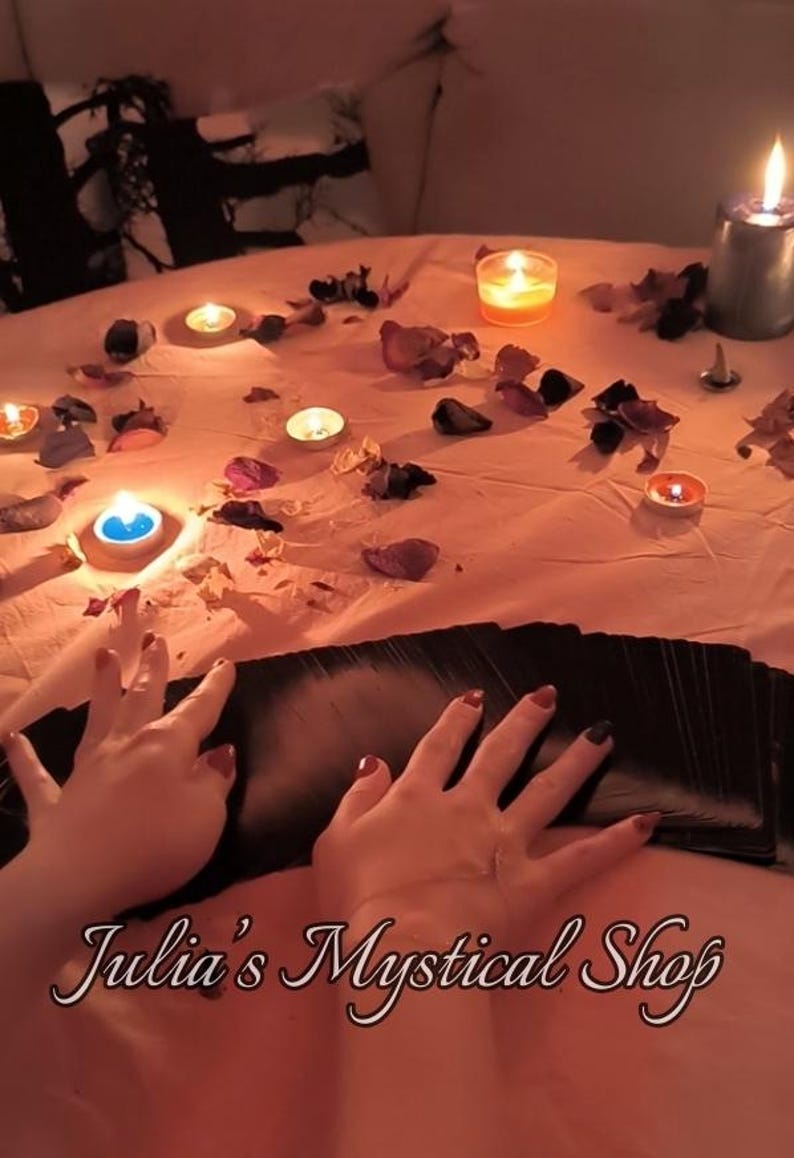 May include: A deck of black tarot cards spread out on a peach-colored surface, surrounded by scattered dried flower petals and several lit tea light candles. The text Julia's Mystical Shop is printed in a white script font at the bottom.