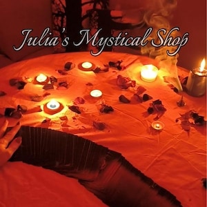 May include: A deck of black tarot cards spread out on a vibrant orange cloth, surrounded by scattered rose petals and several lit tea light candles. The text Julia's Mystical Shop is visible in the upper portion of the image, with wisps of smoke rising.