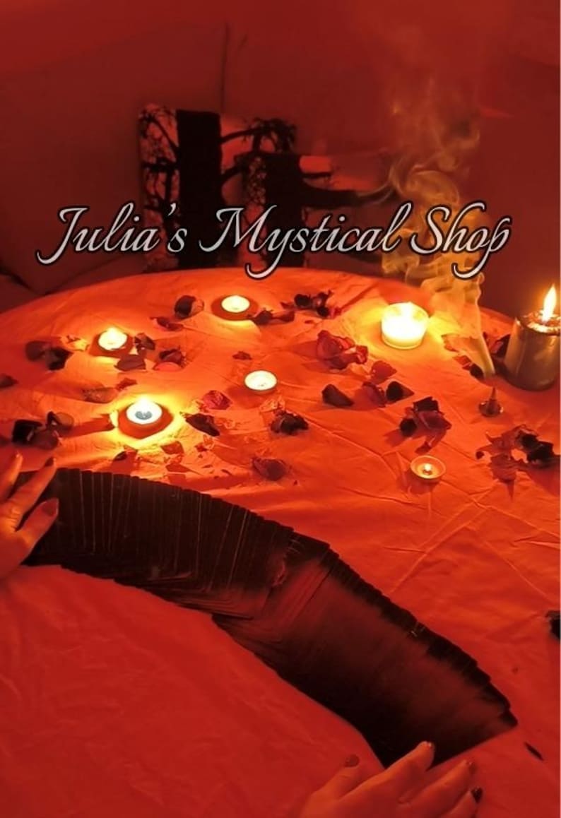 May include: A deck of black tarot cards spread out on a vibrant orange cloth, surrounded by scattered rose petals and several lit tea light candles. The text Julia's Mystical Shop is visible in the upper portion of the image, with wisps of smoke rising.