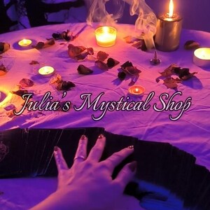 May include: A mystical scene with lit candles, dried petals, and smoke. The text "Julia's Mystical Shop" is displayed. A hand with painted nails hovers over a deck of cards, creating a mysterious atmosphere.