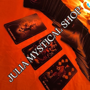 May include: A deck of tarot cards with black backgrounds and dark, detailed illustrations. Three cards are face up, showing The Hanged Man, a group scene, and the Six of Pentacles. The text JULIA MYSTICAL SHOP is overlaid across the cards.