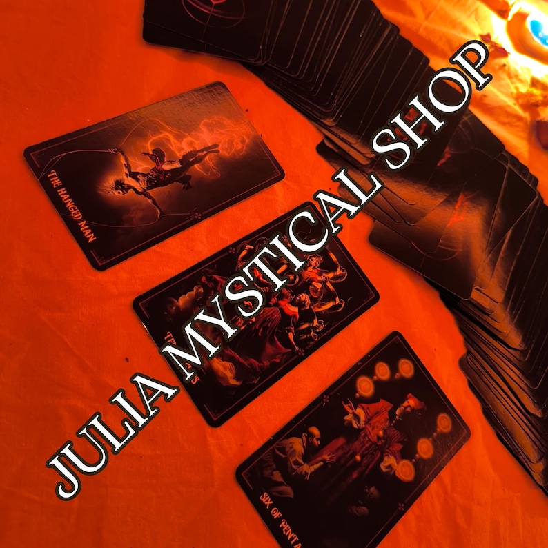 May include: A deck of tarot cards with black backgrounds and dark, detailed illustrations. Three cards are face up, showing The Hanged Man, a group scene, and the Six of Pentacles. The text JULIA MYSTICAL SHOP is overlaid across the cards.