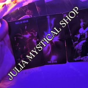 May include: A set of three tarot cards illuminated in vibrant purple light. The cards display the Seven of Cups, The Star, and the Queen of Wands. The text JULIA MYSTICAL SHOP is overlaid diagonally across the cards in a bold, light-colored font.