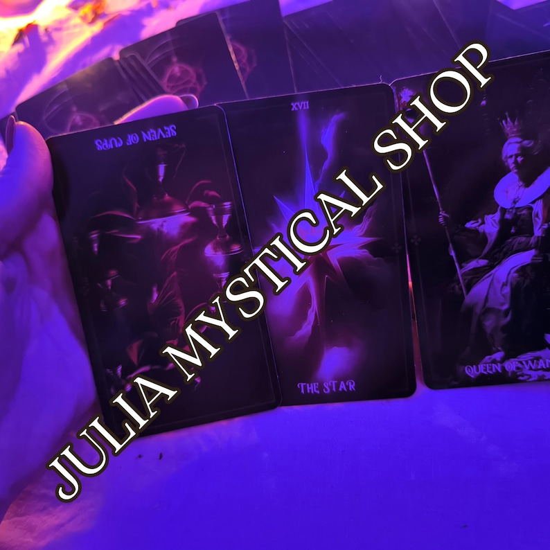 May include: A set of three tarot cards illuminated in vibrant purple light. The cards display the Seven of Cups, The Star, and the Queen of Wands. The text JULIA MYSTICAL SHOP is overlaid diagonally across the cards in a bold, light-colored font.