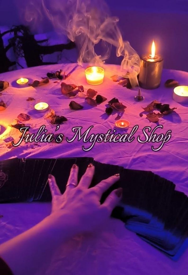 May include: A white tablecloth covered in scattered dried flower petals, multiple lit candles, and a thin trail of incense smoke. A deck of black tarot cards is fanned out in the foreground with the text Julia's Mystical Shop overlaid in white.