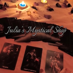 May include: A deck of black tarot cards with the text Julia's Mystical Shop displayed across the center. Three cards are laid out face up, showing The Devil, Knight of Pentacles, and Four of Wands, surrounded by scattered flower petals and lit candles.