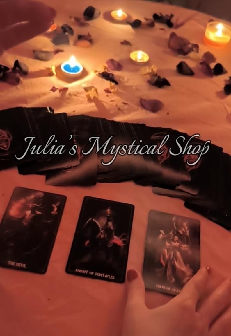 May include: A deck of black tarot cards with the text Julia's Mystical Shop displayed across the center. Three cards are laid out face up, showing The Devil, Knight of Pentacles, and Four of Wands, surrounded by scattered flower petals and lit candles.