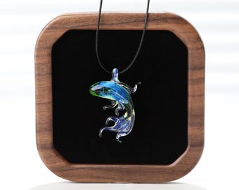 May include: A glass pendant shaped like a swimming fish with swirling blue, green, and clear tones, suspended from a thin black cord. The pendant is displayed against a black square insert within a rounded wooden frame.