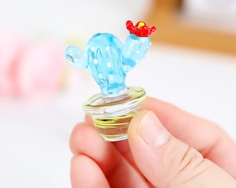 Puede incluir: Una figura de cactus de vidrio en miniatura con un cuerpo azul claro, lunares blancos y una flor roja. El cactus está en una maceta de vidrio transparente. La figura se sostiene en una mano.