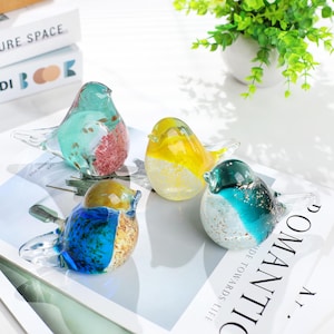 May include: Four colorful glass bird figurines. Each bird has a unique color scheme, including blue, yellow, green, and a mix of colors. The birds are displayed on a white surface, with a plant in the background.