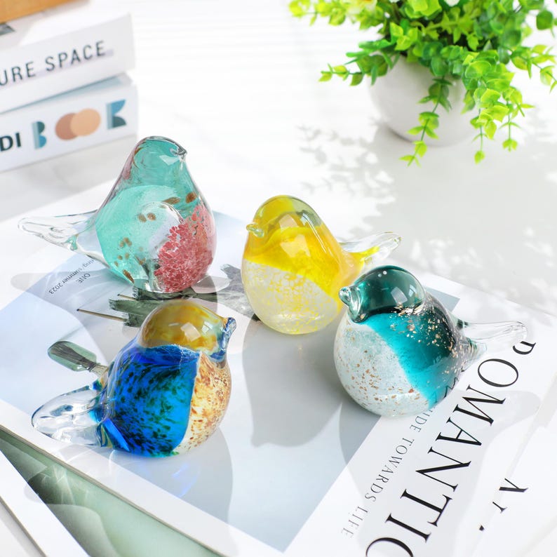 May include: Four colorful glass bird figurines. Each bird has a unique color scheme, including blue, yellow, green, and a mix of colors. The birds are displayed on a white surface, with a plant in the background.