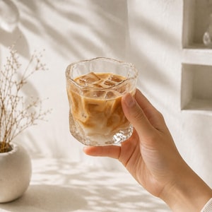 May include: A clear glass tumbler with an irregular, wavy rim and textured, rippled sides. The glass is filled with a light brown iced coffee beverage containing several square ice cubes, showing a gradient of dark coffee and creamy milk.