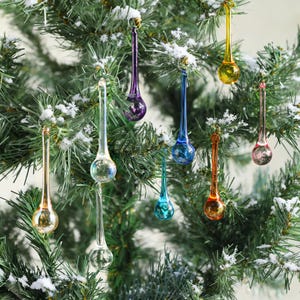 May include: A collection of colorful, teardrop-shaped Christmas ornaments hanging on a snow-covered evergreen tree. The ornaments are in various colors, including gold, purple, blue, and clear, with a long, slender stem and a rounded bulb at the bottom.