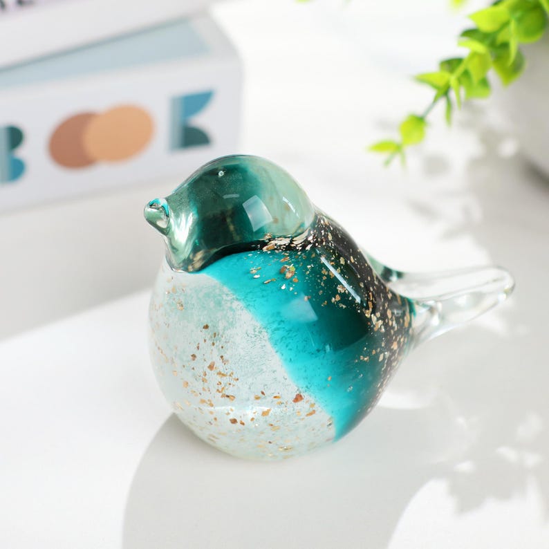 May include: A decorative glass bird figurine. The bird features a teal, white, and black color scheme with gold flecks. The glass bird has a rounded body and a small beak. The figurine is a decorative item.