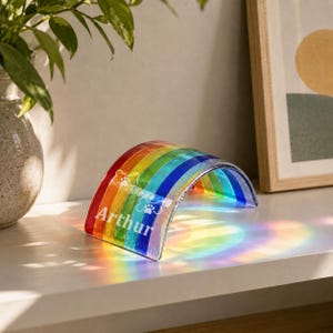 May include: A curved, arched glass ornament featuring a vibrant rainbow spectrum of red, orange, yellow, green, blue, and violet stripes. The piece is personalized with the white text Arthur and two white paw print icons on the surface.