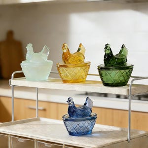 May include: Four glass hen-shaped trinket boxes in pastel colours: light blue, yellow, green, and blue. Each hen sits atop a diamond-patterned bowl. The boxes are displayed on a two-tiered metal shelf, showcasing their decorative design.