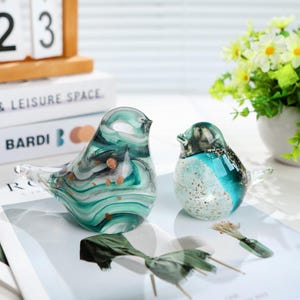 May include: Two glass bird figurines. The larger bird has a teal, white, and black swirl pattern with gold flecks. The smaller bird is teal and white with gold accents. Both have clear beaks and tails. Decorative home decor.