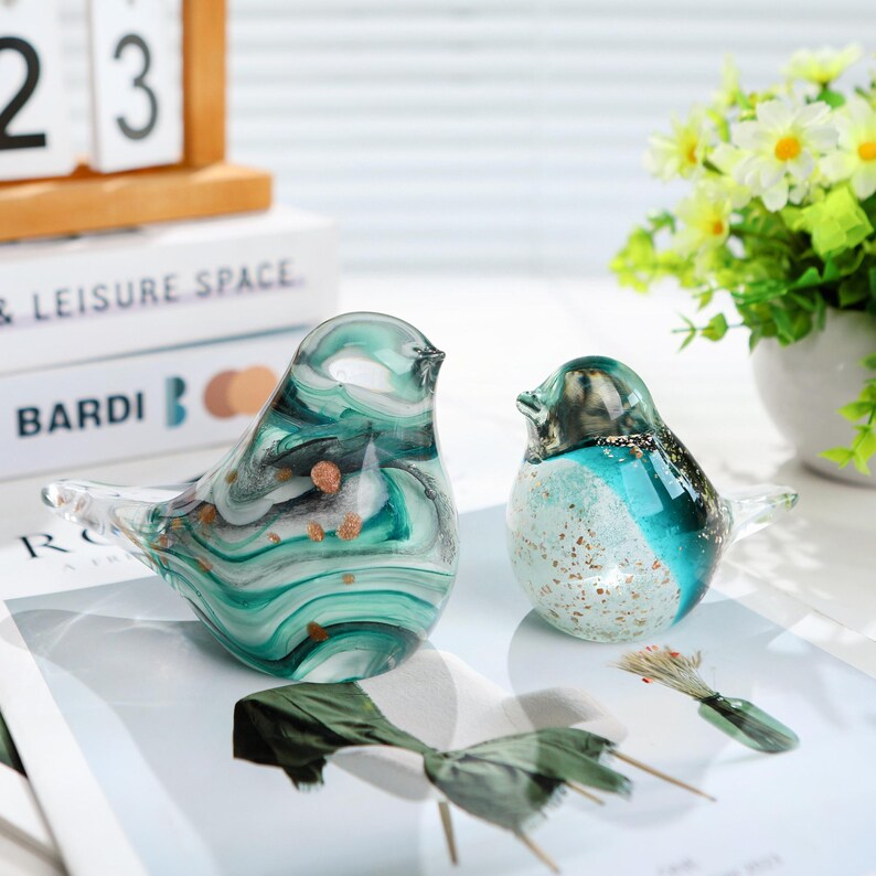 May include: Two glass bird figurines. The larger bird has a teal, white, and black swirl pattern with gold flecks. The smaller bird is teal and white with gold accents. Both have clear beaks and tails. Decorative home decor.