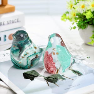 May include: Two decorative glass bird figurines. One bird is teal and white with gold accents, the other is teal, red, and white with gold accents. The birds are on a white surface with a magazine and a small green decoration.