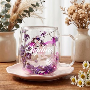 Op de afbeelding: Een heldere glazen mok met een handvat, gevuld met paarse en roze gedroogde bloemen, en de naam "Sophia" in witte letters. De mok staat op een lichtroze schotel. Decoratieve bloemen staan op de achtergrond.
