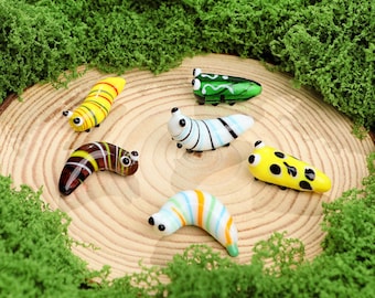 May include: A collection of six colorful glass caterpillar figurines. The caterpillars are in various colors, including yellow, green, blue, and brown, with striped and spotted patterns. They are arranged on a wooden slice surrounded by green moss.
