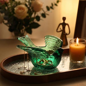 Op de afbeelding: Een decoratieve, smaragdgroene glazen vogelvormige sieradenschaal. De schaal bevat gouden kettingen en ringen. Een brandende kaars en een klein bronzen beeldje staan op de achtergrond. De schaal staat op een houten dienblad.