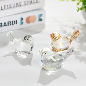 May include: Three decorative glass bird figurines. One is clear, one is amber, and one is iridescent. The birds have detailed wings and beaks, and are displayed on a white surface. The birds are a decorative item.