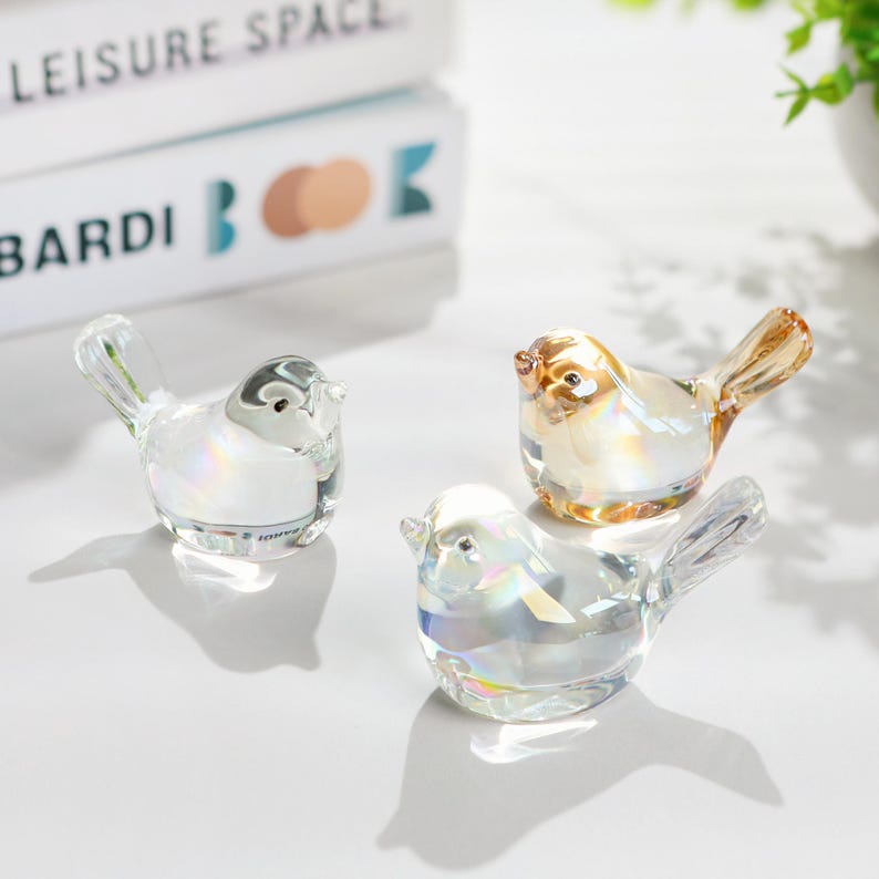 May include: Three decorative glass bird figurines. One is clear, one is amber, and one is iridescent. The birds have detailed wings and beaks, and are displayed on a white surface. The birds are a decorative item.