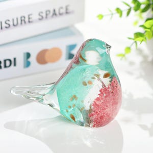 May include: A decorative glass bird figurine with a clear, turquoise, and red design. The bird has a transparent tail and wings, with gold flecks throughout the body. The figurine is a decorative item.