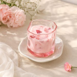 May include: A clear, wavy-rimmed glass tumbler filled with a creamy pink strawberry milk beverage containing visible red strawberry pieces. The glass sits on a small, irregular white ceramic saucer, accompanied by soft pink flowers.