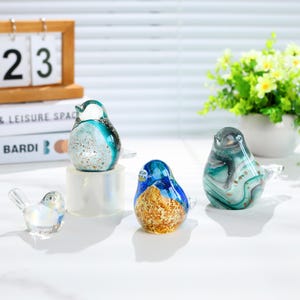May include: A collection of colorful glass bird figurines. The birds feature various designs, including blue, green, and gold, with iridescent and speckled details. The figurines are displayed on a white surface, with a calendar and flowers in the background.