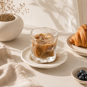 May include: A clear, rippled glass tumbler filled with iced coffee, resting on a matching off-white ceramic saucer. The glass features an irregular, organic shape with visible ice cubes submerged in the light brown coffee liquid.