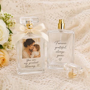 May include: Two clear glass perfume bottles with gold spray nozzles. One bottle features a photo of a person holding a child with the text "You are my greatest blessing." and a cream ribbon. The other bottle displays the text "Forever grateful, always yours."