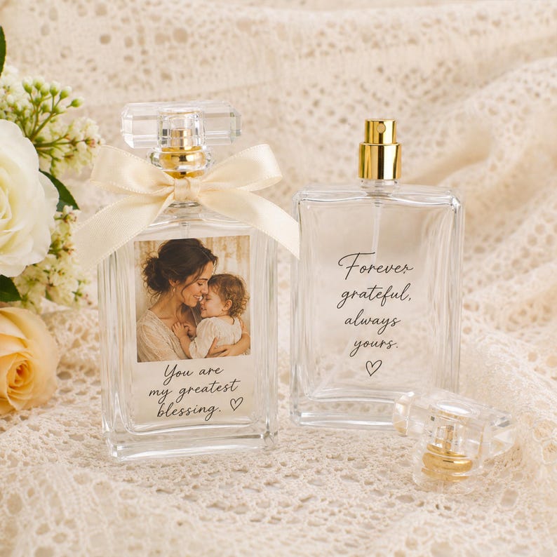 May include: Two clear glass perfume bottles with gold spray nozzles. One bottle features a photo of a person holding a child with the text "You are my greatest blessing." and a cream ribbon. The other bottle displays the text "Forever grateful, always yours."