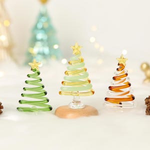 Puede incluir: Tres árboles de Navidad decorativos de cristal en verde, amarillo y ámbar. Cada árbol tiene un diseño en espiral y una estrella dorada en la parte superior. Los árboles están sobre una superficie blanca con un fondo borroso.