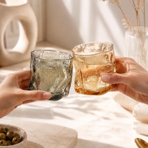 May include: Two short drinking glasses with an irregular, rippled texture. One glass is tinted in a smoky grey hue, while the other features a warm amber tone. Both vessels have a thick base and a wavy, organic rim, reflecting light through the glass.