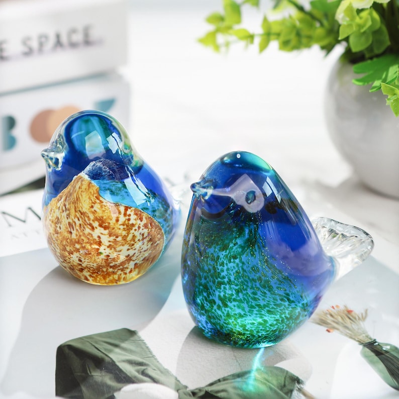 May include: Two decorative glass bird figurines. One bird features a blue and gold body, the other a blue and green body. The birds have clear glass wings and beaks. The figurines are sitting on a white surface.