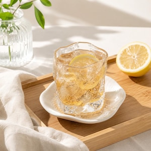May include: A clear, textured glass tumbler filled with a golden liquid, ice cubes, and a lemon slice, resting on a white, wavy-edged ceramic saucer. The glass features an irregular, organic shape with a rippled surface texture throughout.
