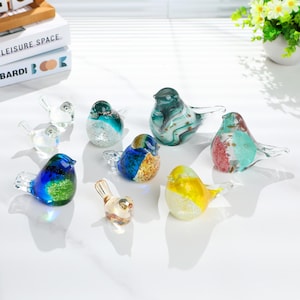 May include: A collection of colorful glass bird figurines. The birds feature various colors, including blue, green, yellow, and red, with clear glass wings. The figurines are arranged on a white surface, showcasing their decorative nature.