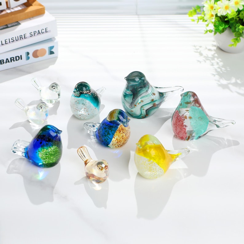 May include: A collection of colorful glass bird figurines. The birds feature various colors, including blue, green, yellow, and red, with clear glass wings. The figurines are arranged on a white surface, showcasing their decorative nature.