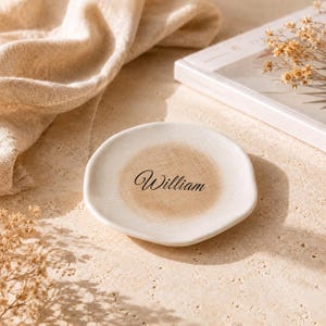 May include: A round, off-white ceramic trinket dish with an irregular, organic edge. The center of the dish features a soft, tan-colored circular wash and the name William printed in a black, elegant, cursive script font.