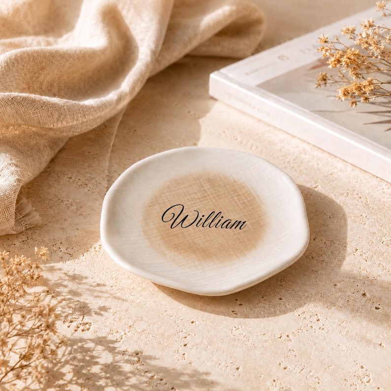 May include: A round, off-white ceramic trinket dish with an irregular, organic edge. The center of the dish features a soft, tan-colored circular wash and the name William printed in a black, elegant, cursive script font.