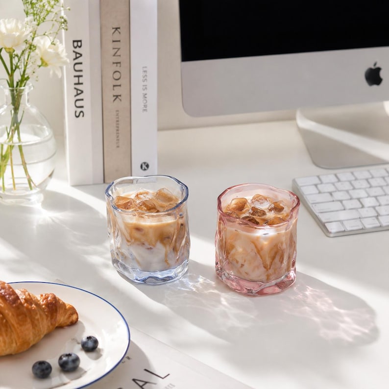 May include: Two short, textured glass tumblers filled with iced coffee. One glass has a subtle blue tint, while the other has a soft pink tint. Both glasses feature an irregular, wavy rim and a rippled surface, containing ice cubes and coffee.