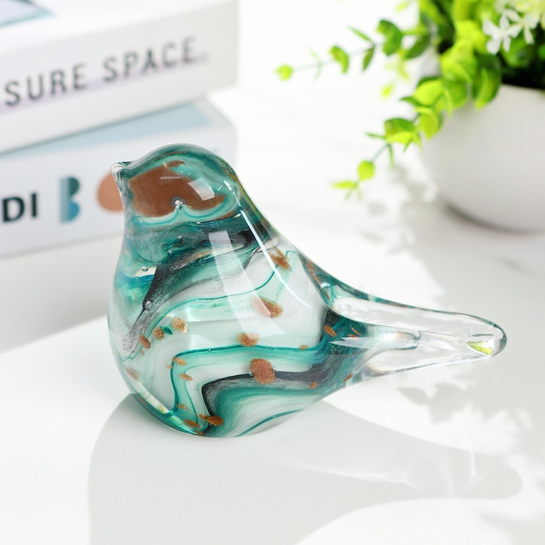 May include: A decorative glass bird figurine. The bird features a teal, white, and black swirling design with gold flecks. The figurine is perched on a white surface. The bird's beak and head are brown.