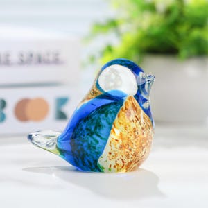 May include: A decorative glass bird figurine. The bird features a rounded body with a blue, white, and amber color scheme. The glass is translucent, allowing light to pass through. The bird is perched on a white surface.