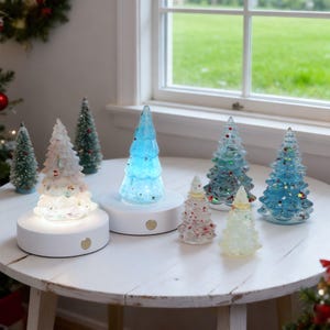 May include: A collection of decorative Christmas tree figurines in various sizes and colors. Some trees are clear with colorful accents, while others have a blue or white glow. The trees are displayed on a white table near a window.