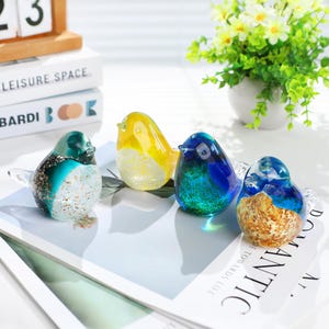 May include: Four colorful glass bird figurines. The birds are in shades of yellow, blue, teal, and brown, with clear glass wings. They are arranged on a white surface with text. A small white vase with green and white flowers is in the background.