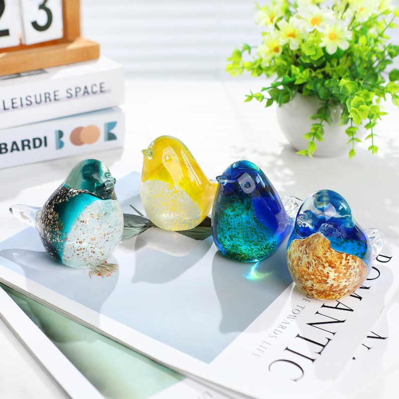 May include: Four colorful glass bird figurines. The birds are in shades of yellow, blue, teal, and brown, with clear glass wings. They are arranged on a white surface with text. A small white vase with green and white flowers is in the background.